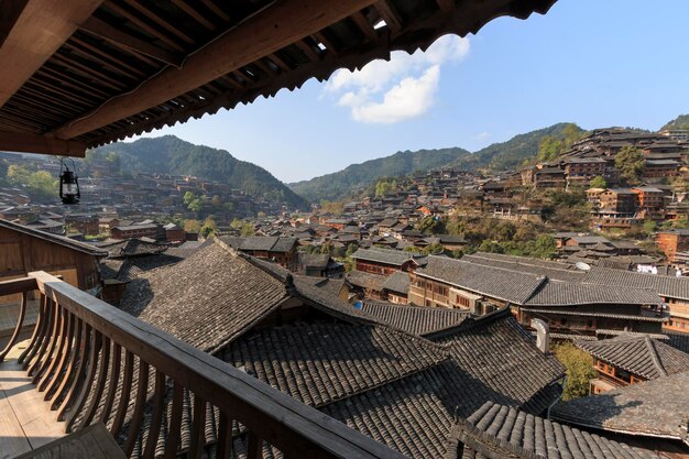 A panoramic view of Lijiang Ancient Town, showing the traditional Naxi architecture with tiled roofs and wooden structures. A clear blue river runs through the town, with weeping willow trees lining its banks. In the background, the snow-capped Jade Dragon Snow Mountain is visible.